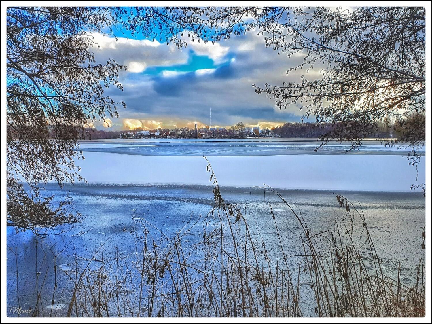 Bildbeschreibung: 
Blick durch Zweige und Äste auf den noch teilweise mit Eis und Schnee bedeckten Ostorfer See. Graue und blaue Wolken ziehen am Himmel vorbei. In weiterer Ferne sind der Fernsehturm und einige Gebäude zu sehen, die durch Sonnenstrahlen in einer Wolkenlücke in goldgelbes Licht getaucht werden.
